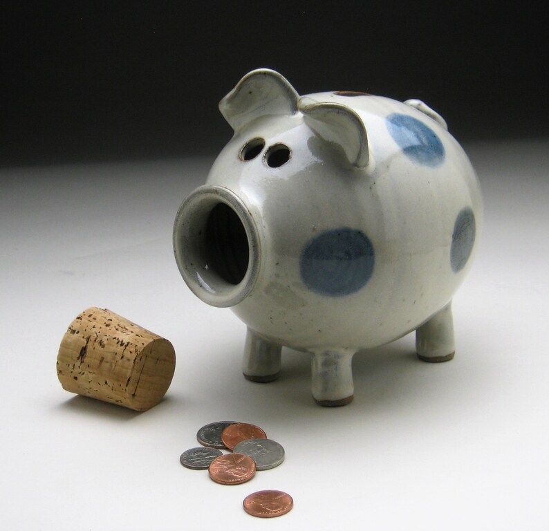 K&ouml;nnte beinhalten: Ein wei&szlig;es Keramik-Sparschwein mit blauen Punkten und einem Korkstopfen. Das Sparschwein hat vier Beine und eine Schnauze. Vor dem Sparschwein liegen mehrere M&uuml;nzen auf dem Tisch.