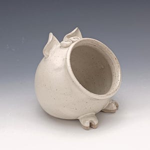 Pode incluir: Saleiro de cerâmica branca com um esmalte salpicado. O porco tem uma grande abertura para despejar sal e duas orelhas pequenas.