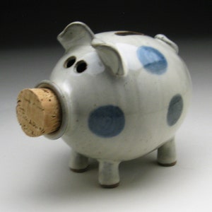 K&ouml;nnte beinhalten: Ein wei&szlig;es Keramik-Sparschwein mit blauen Punkten und einem Korkstopfen f&uuml;r den M&uuml;nzeinschub.