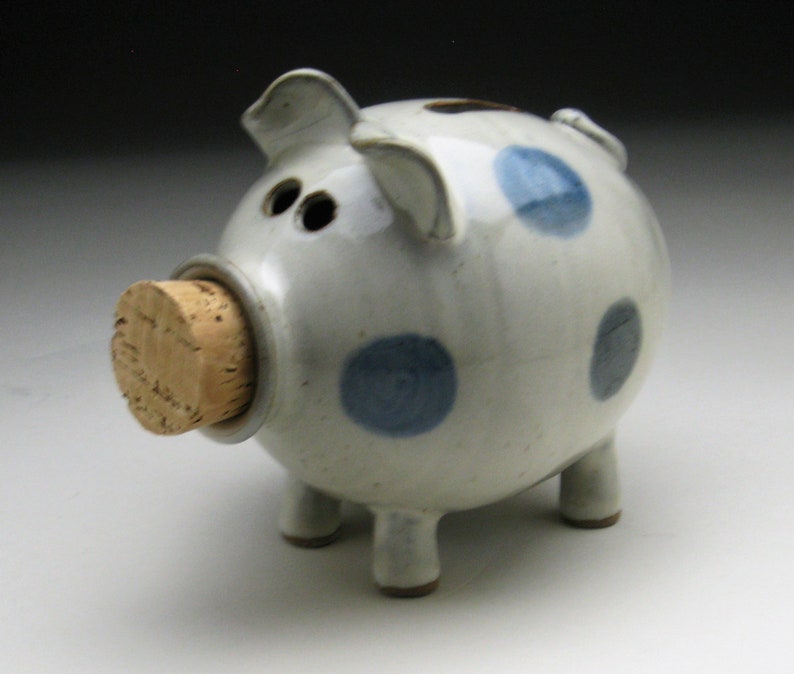 K&ouml;nnte beinhalten: Ein wei&szlig;es Keramik-Sparschwein mit blauen Punkten und einem Korkstopfen f&uuml;r den M&uuml;nzeinschub.