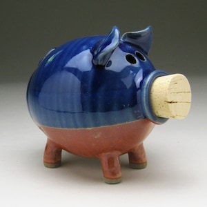 May include: A blue and brown ceramic piggy bank with a cork stopper in the snout. The piggy bank has a glossy finish and is decorated with a blue and brown glaze.