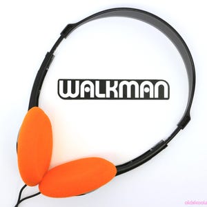 May include: Overhead shot of vintage Walkman headphones. The headphones have a black headband and ear pieces with bright orange foam pads. The word "WALKMAN" is printed in white block letters on the white background.