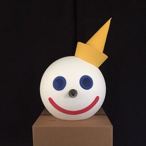 May include: A white, round, smiling face mask with blue eyes and a red mouth. The mask has a yellow cone-shaped hat on top. The mask is sitting on a brown cardboard box.