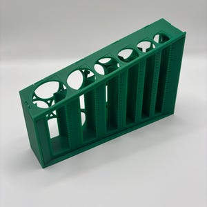 Puede incluir: Un soporte de plástico verde para tubos de ensayo con siete aberturas circulares y divisores verticales. El soporte está inclinado, con marcas de medición a lo largo de los divisores. Diseñado para contener tubos de ensayo para uso científico o de laboratorio.