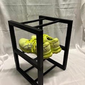 May include: A pair of neon yellow athletic shoes rest inside a black, geometric shoe rack. The shoes have gray accents and are positioned to showcase the soles. The rack is made of square metal tubing and has a modern design.
