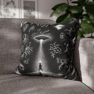 Puede incluir: Cojín decorativo en blanco y negro con una ilustración surrealista de un OVNI que proyecta un haz de luz sobre una silueta. El diseño incluye varios dibujos de caras y formas abstractas.