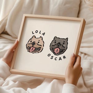 Op de afbeelding: Een ingelijste print met cartoon illustraties van honden. Eén hond is bruin, de andere grijs, beide met hun tong uit. De namen "LOLA" en "OSCAR" staan boven en onder de honden. De lijst is van lichtgekleurd hout.