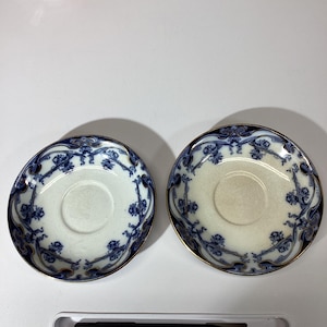 May include: Two round ceramic saucers featuring a white center and a decorative border with dark blue floral patterns and gold trim. The saucers have a circular indentation in the middle for a cup, with one saucer appearing slightly darker in tone.