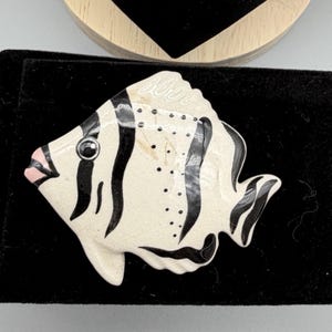 May include: A ceramic fish-shaped brooch with a cream-colored base and black stripes. The fish has a pink mouth and a black eye. The brooch is on a black surface.