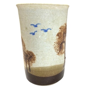 May include: A ceramic tumbler with a landscape design. The off-white tumbler features brown trees and a brown base. Blue birds fly across the top. The tumbler is approximately 5 inches tall.