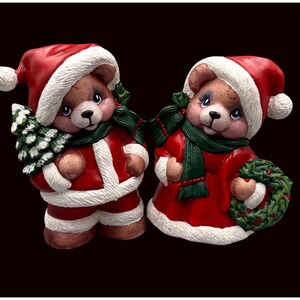 May include: Two ceramic Christmas bear figurines. One bear wears a red Santa suit and hat, holding a small green Christmas tree. The other bear wears a red dress and hat, holding a green wreath. Both bears have green scarves.