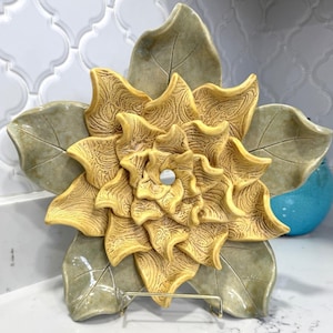 Könnte beinhalten: Dekorative Keramikschale in Blumenform mit einem zentralen Loch. Die Schale hat ein strukturiertes, geschichtetes Design in Gelb- und Beigetönen mit großen, olivgrünen Blattakzenten. Sie wird von einem goldfarbenen Metallständer getragen.