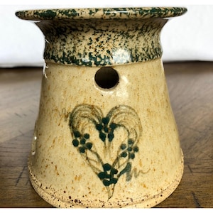 Könnte beinhalten: Ein beigefarbener Keramik-Ölwärmer mit gesprenkelter Oberfläche und dunkelgrünem Rand. Der Körper zeigt ein handgemaltes dunkelgrünes Herzmotiv mit floralen Akzenten und ein kreisförmiges Belüftungsloch nahe der Oberseite.
