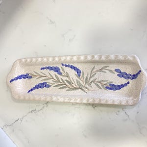 Könnte beinhalten: Ein rechteckiges Keramiktablett mit gesprenkelter cremeweißer Oberfläche und gewelltem Rand. In der Mitte befindet sich ein handgemaltes botanisches Motiv aus blauen Lavendelzweigen mit grünen Blättern, die horizontal angeordnet sind.