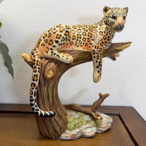 May include: A ceramic figurine of a leopard resting on a tree branch. The leopard has a cream-colored coat with black and orange spots. The tree trunk is brown with textured bark, and the base features green moss-like details and a light gray rock.