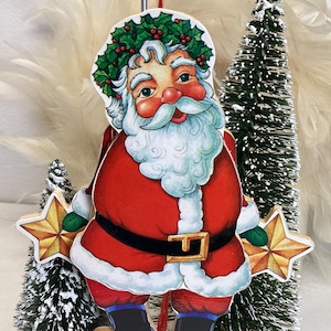 May include: A flat, die-cut wooden Santa Claus ornament featuring a red suit with white trim, a black belt with a gold buckle, and a green holly wreath on the head. Santa holds a golden yellow star in each hand, with a white beard and rosy cheeks.
