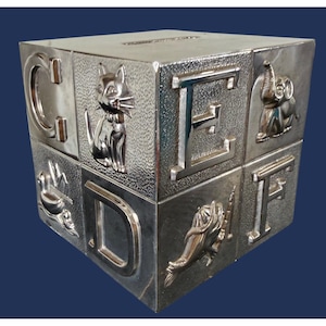 May include: A silver-toned metallic cube-shaped coin bank featuring raised relief designs on its sides. The faces display the letters C, D, E, and F, alongside embossed figures of a cat, a bird, an elephant, and a fish. A coin slot is visible on the top surface.