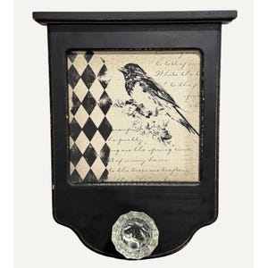 Könnte beinhalten: Schwarzes, an der Wand montiertes dekoratives Regal mit einem Used-Look-Finish. Das Regal zeigt einen gerahmten Druck eines Vogels und Text, mit einem Harlekinmuster auf der linken Seite. Ein klarer, facettierter Knopf befindet sich unten.