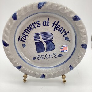 May include: A round, stoneware plate with a gray base and blue accents. The plate features the text "Farmers at Heart" and "BECK'S" in blue, along with a stylized "B" design. A sticker with the American flag is visible.