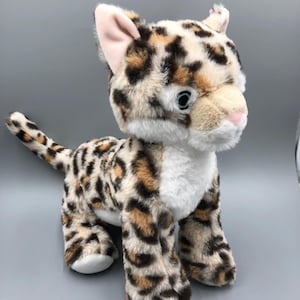 May include: A plush leopard toy with a spotted pattern in shades of brown, tan, and black. The toy has a white chest and face, pink inner ears, and blue eyes. The leopard is standing on all fours with its tail extended.