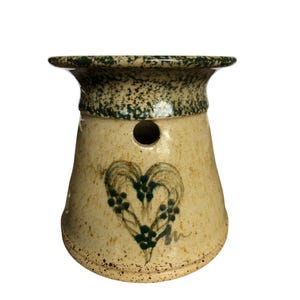 May include: A ceramic oil warmer with a heart design. The warmer is beige with a green speckled rim and a heart painted in green. It has a hole for ventilation and a flared top.