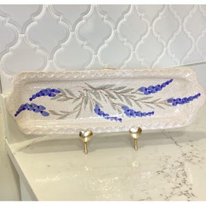 May include: A rectangular ceramic serving tray with a speckled cream-colored finish and scalloped edges. The center features a hand-painted botanical design of blue lavender sprigs and sage green leaves. The tray is displayed on a gold metal stand.