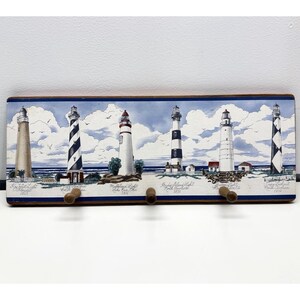 May include: A rectangular wooden wall hook rack featuring a painted scene of six distinct lighthouses against a cloudy sky and ocean backdrop. The lighthouses are labeled with their names and locations, and three wooden pegs are attached to the bottom.