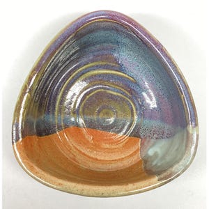 Może przedstawiać: Ręcznie robiona ceramiczna miska o unikalnym trójkątnym kształcie. Misa ma mieszankę kolorów, w tym pomarańczowy, niebieski, fioletowy i zielony. Wnętrze ma spiralny wzór, a zewnętrzna strona ma teksturowane wykończenie. Ta dekoracyjna miska jest idealna do serwowania przekąsek lub jako element dekoracyjny.