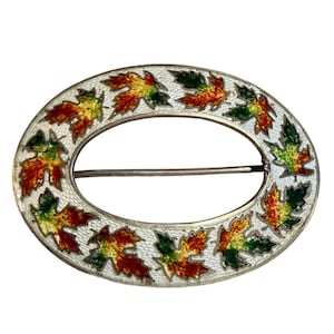 May include: An oval-shaped brooch featuring a silver-toned metal frame with a textured white background. The piece is decorated with a repeating pattern of enameled maple leaves in vibrant shades of red, orange, yellow, and green.