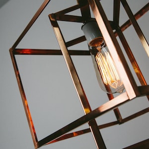 Puede incluir: Lámpara colgante geométrica de color cobre con una bombilla Edison visible. La lámpara tiene un diseño en forma de cubo con barras de metal que se cruzan. La bombilla emite un brillo cálido y anaranjado, que contrasta con el fondo gris frío.