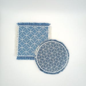 May include: A square denim coaster with a white circular geometric pattern and a round denim coaster with a white isometric cube pattern. Both coasters feature frayed edges and white stitched borders, showcasing a blue fabric texture throughout.