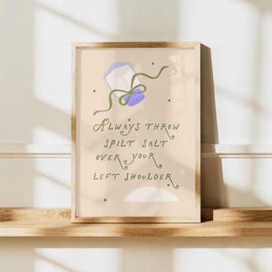 Könnte beinhalten: Ein rechteckiger Wandkunstdruck auf hellbeigem Hintergrund mit einem violetten Salzstreuer, um den ein grünes Band gebunden ist. Der Text lautet Always throw spilt salt over your left shoulder, ergänzt durch kleine Sternillustrationen und einen weißen Salzhaufen am unteren Rand.