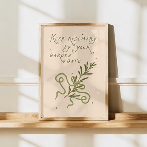 Puede incluir: Una impresión vertical enmarcada sobre un fondo beige claro que presenta una ilustración de una rama de romero verde con líneas onduladas y pequeñas estrellas. El texto dice: Keep rosemary by your garden gate. Marco de madera clara.
