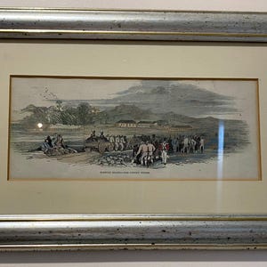 May include: Framed antique print showing a historical scene with figures, buildings, and a mountain backdrop. The print is in a silver-toned frame. Text at the bottom of the print is partially visible.