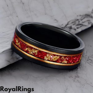 Puede incluir: Un anillo de carburo de tungsteno negro con una incrustación roja y dorada. La incrustación presenta un patrón de motas rojas y doradas.