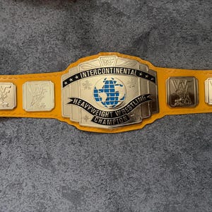 May include: A yellow wrestling championship belt featuring a large central gold-tone metal plate with the text INTERCONTINENTAL HEAVYWEIGHT WRESTLING CHAMPION and a blue globe graphic. Four smaller square gold-tone metal plates with WWF logos.