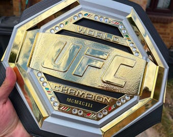 Può includere: Una cintura da campione del mondo UFC ottagonale con una grande placca centrale color oro con il testo UFC e CHAMPION, il numero romano MCMXCIII e un bordo di cristalli trasparenti e piccoli accenti rossi, bianchi e verdi su una cinghia nera.