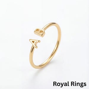 Op de afbeelding: Een goudkleurige ring met de letters "A" en "B" in een open ontwerp. De ring staat tegen een witte achtergrond. De tekst "Royal Rings" staat onderaan de afbeelding.