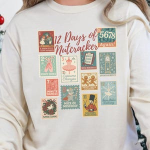 May include: A cream-colored long-sleeve sweatshirt with a colorful graphic of postage stamps that read "12 Days of Nutcracker Again!". The person is wearing red and black plaid pajama pants and reindeer slippers. Christmas trees and decorations are in the background.