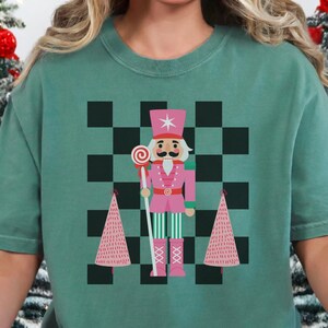 May include: A green t-shirt featuring a pink nutcracker soldier design with a checkerboard background and two pink Christmas trees. The person is wearing red and black plaid pajama pants. Christmas trees and decorations are in the background.