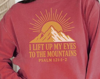 May include: A maroon sweatshirt with a graphic of a mountain and sun design, with the text "I lift up my eyes to the mountains" and Psalm 121:1-2. The sweatshirt is worn with blue jeans.