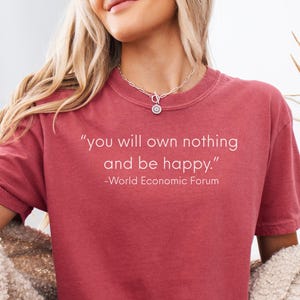 May include: A dusty rose-colored t-shirt with the quote "you will own nothing and be happy." printed in white. The text is from the World Economic Forum. The model is wearing denim shorts and a cream-colored cardigan.