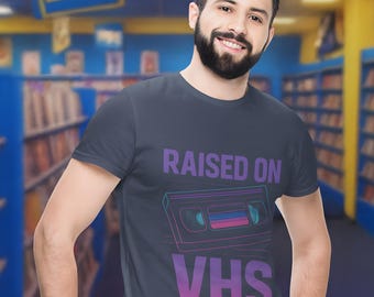 May include: Dark gray t-shirt with a retro VHS tape graphic and the text "RAISED ON VHS" in purple and pink. The person is wearing blue jeans. The background is a video rental store.