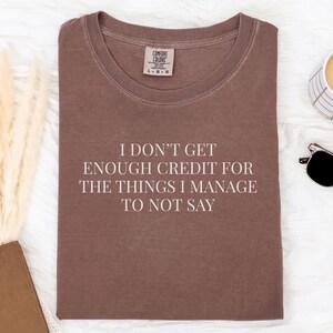 May include: A brown t-shirt with the text "I DON'T GET ENOUGH CREDIT FOR THE THINGS I MANAGE TO NOT SAY". The shirt is laid flat on a white surface, with a cup of coffee, sunglasses, and a notebook nearby.