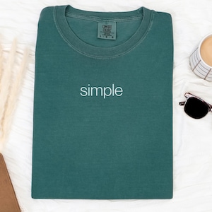May include: A teal-colored t-shirt with the word "simple" in white lettering. The shirt is laid flat on a white surface, next to a cup of coffee, sunglasses, and a brown notebook.