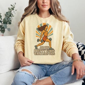 May include: A light yellow sweatshirt with a retro graphic of a woman lifting weights, with the text "ALL THE PRETTY GIRLS WALK LIKE THIS". The design features orange, brown, and beige colors. The sweatshirt has long sleeves.