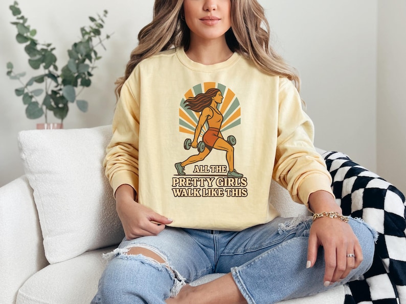 May include: A light yellow sweatshirt with a retro graphic of a woman lifting weights, with the text "ALL THE PRETTY GIRLS WALK LIKE THIS". The design features orange, brown, and beige colors. The sweatshirt has long sleeves.