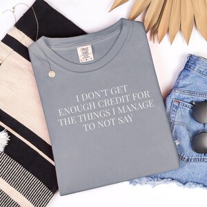 May include: A gray t-shirt with the text "I DON'T GET ENOUGH CREDIT FOR THE THINGS I MANAGE TO NOT SAY" is displayed with denim shorts, sunglasses, a tote bag, and a necklace.