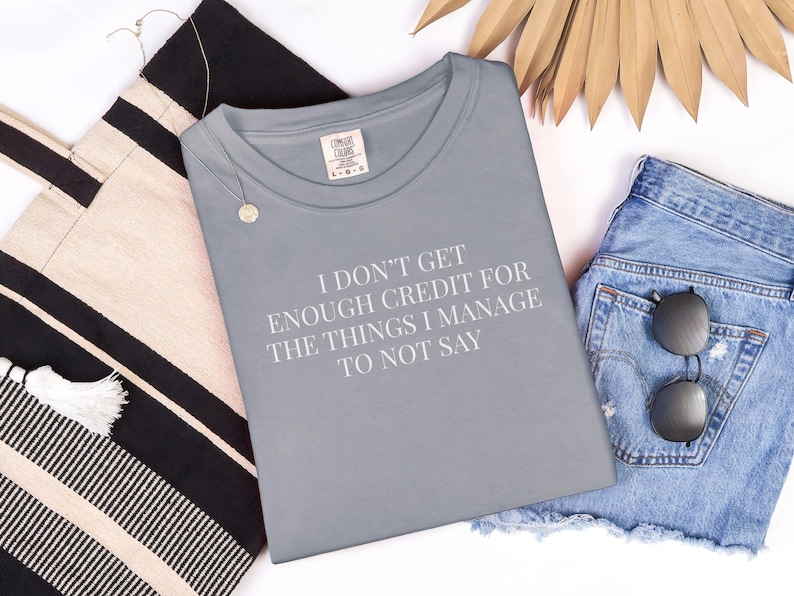 May include: A gray t-shirt with the text "I DON'T GET ENOUGH CREDIT FOR THE THINGS I MANAGE TO NOT SAY" is displayed with denim shorts, sunglasses, a tote bag, and a necklace.