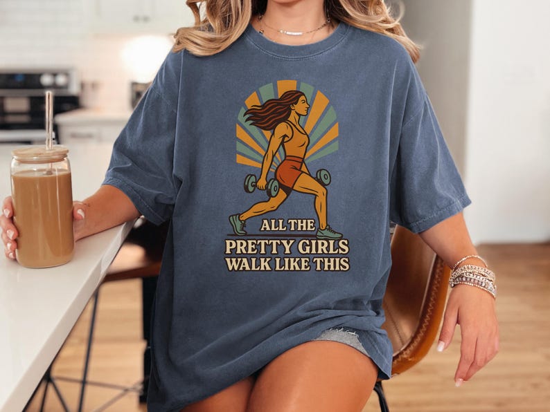 May include: Blue t-shirt with a graphic of a woman lifting weights, set against a retro sunburst design. The text "ALL THE PRETTY GIRLS WALK LIKE THIS" is printed below the image. The shirt is a comfortable, casual style.